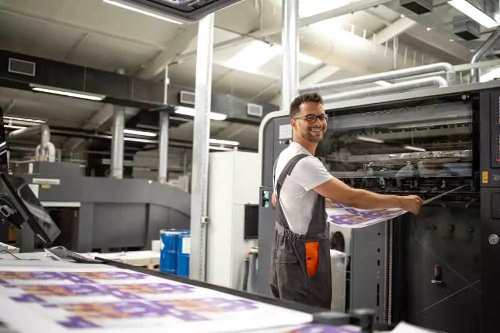 Hombre sonriente con gafas operando una gran máquina de impresión industrial y revisando una hoja impresa, ilustrando un ejemplo de las oportunidades de Empleo Packaging en el sector gráfico.